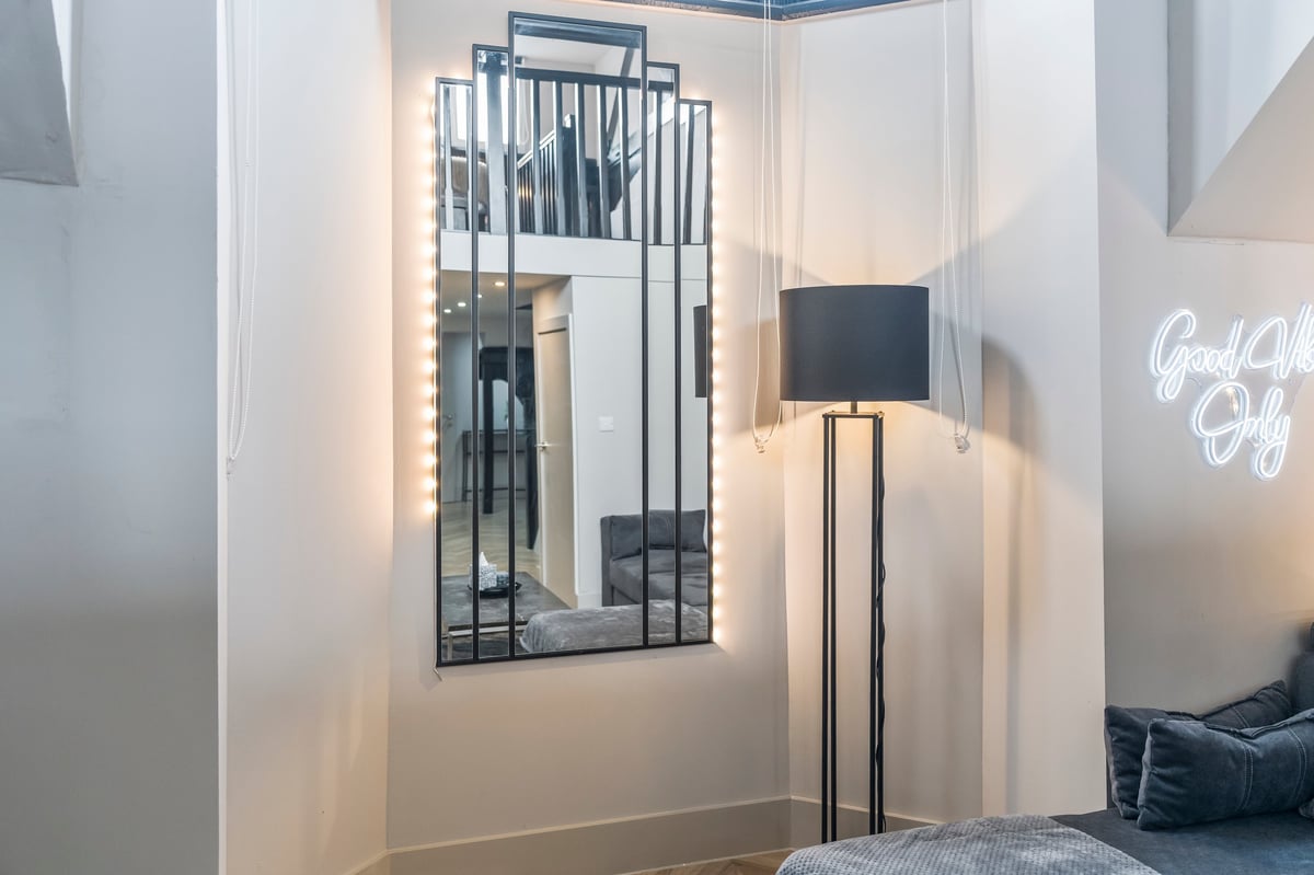 Bedroom corner at Athenaeum House with a black-framed mirror, floor lamp and a neon sign on the white walls