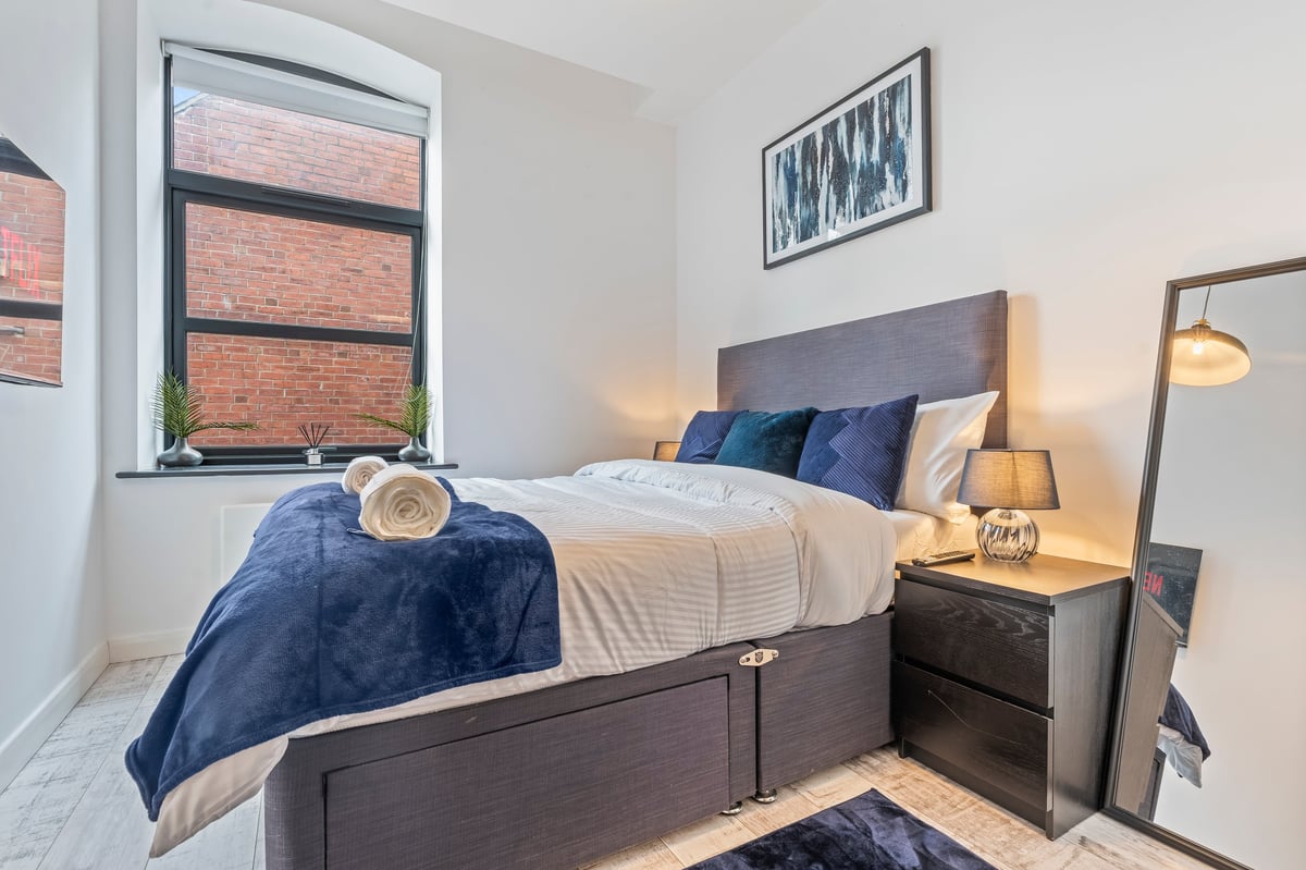 Bedroom at The Calls with a grey bed, blue bedding, gold lamps and a brick accent wall with a large window