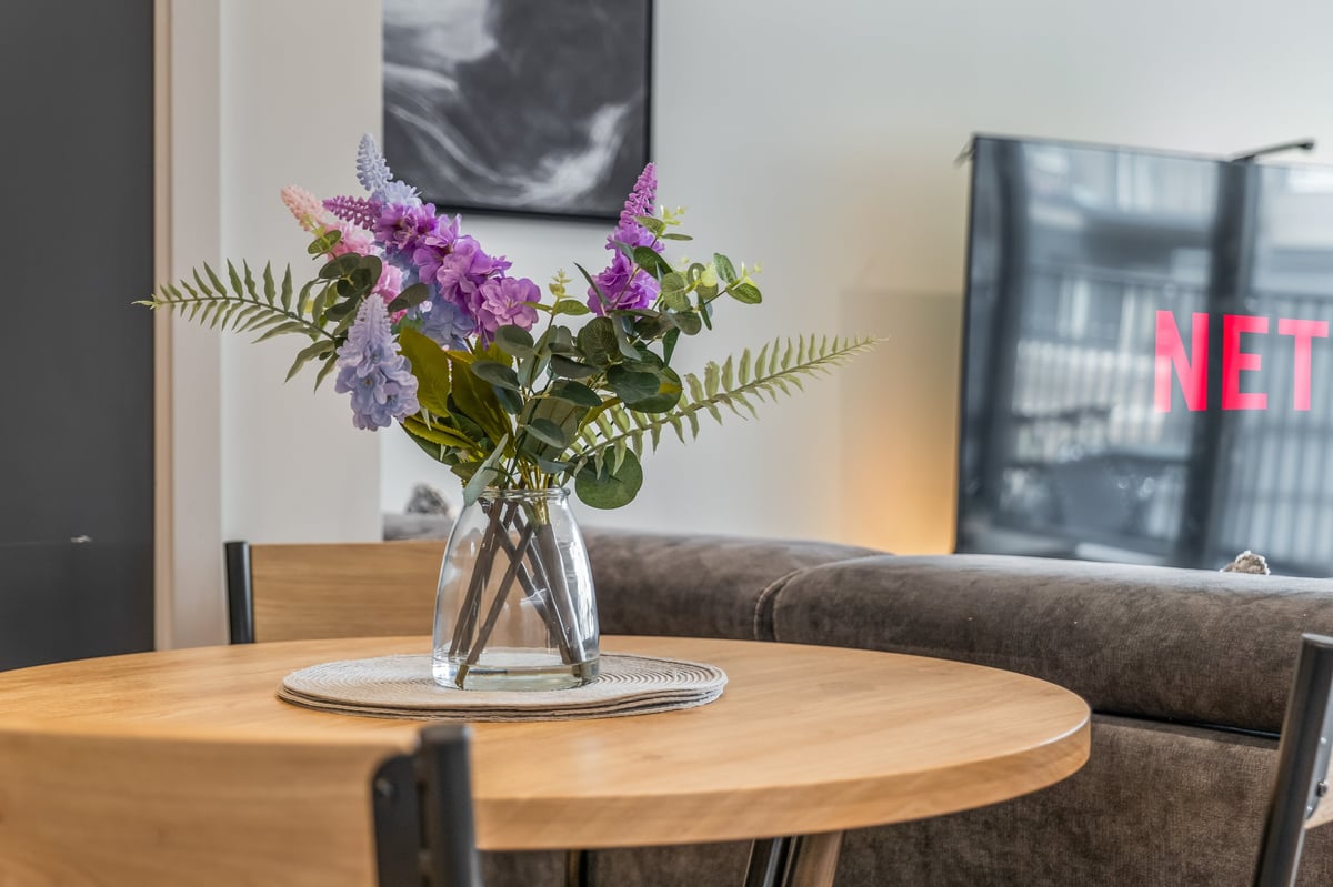 Glass vase of purple and white flowers with green foliage on a round wooden dining table at the Aspire apartment, with the modern living room behind