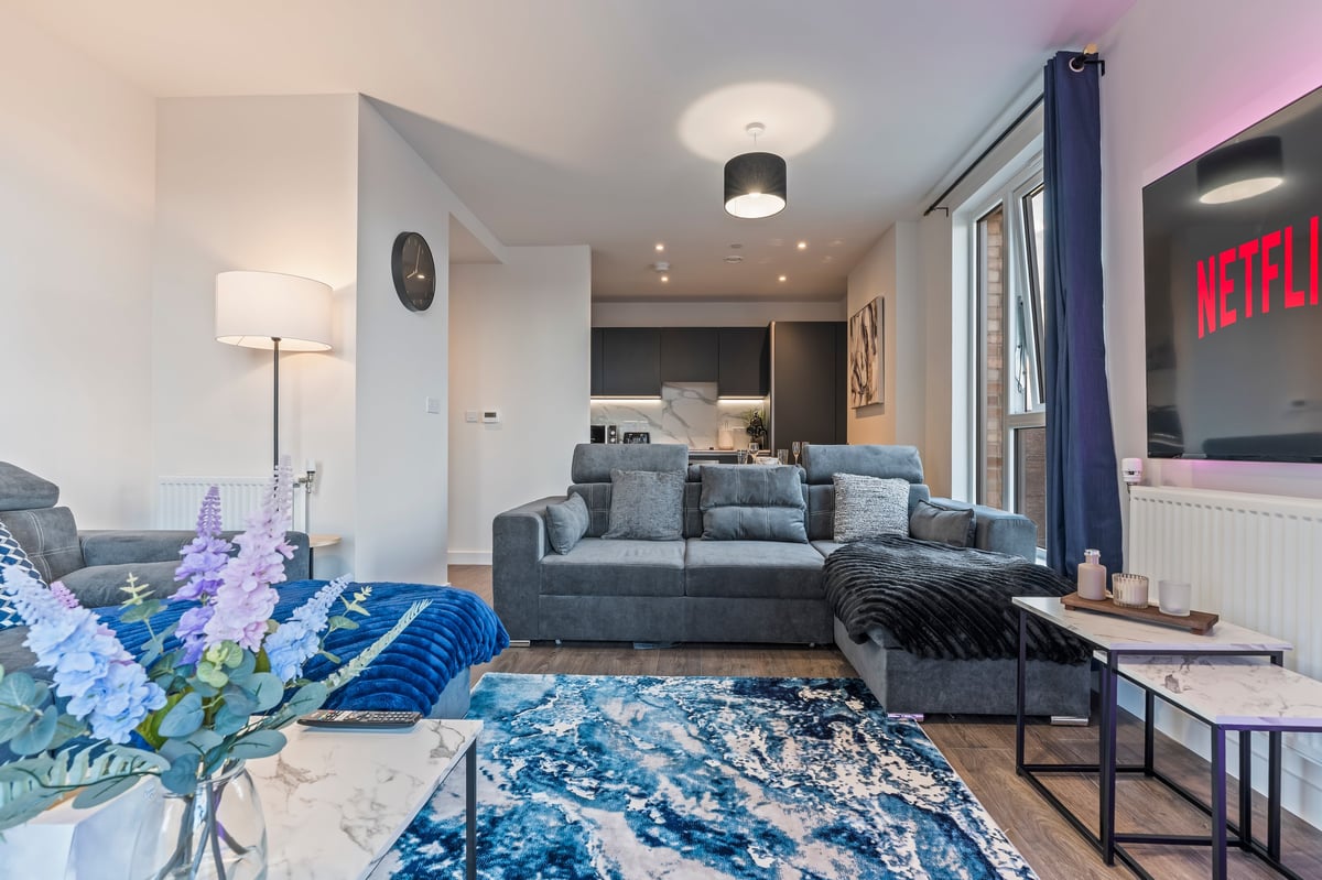Open-plan living space at Wedgewood with a grey sofa, blue patterned area rug, wall-mounted television and contemporary furnishings leading into the kitchen
