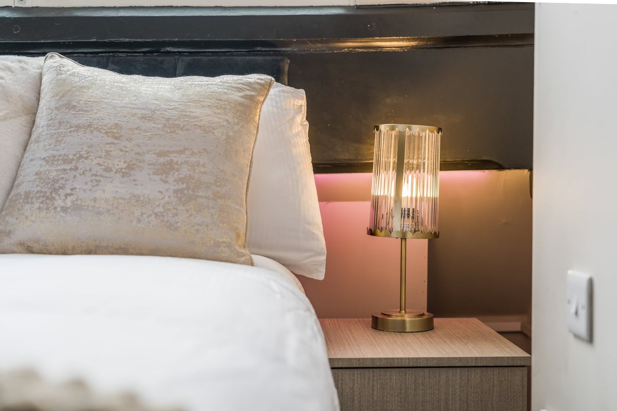 Bedroom corner with white pillows on the bed, a decorative lamp with brass base on the nightstand and warm ambient lighting against the terracotta accent wall at Athenaeum House