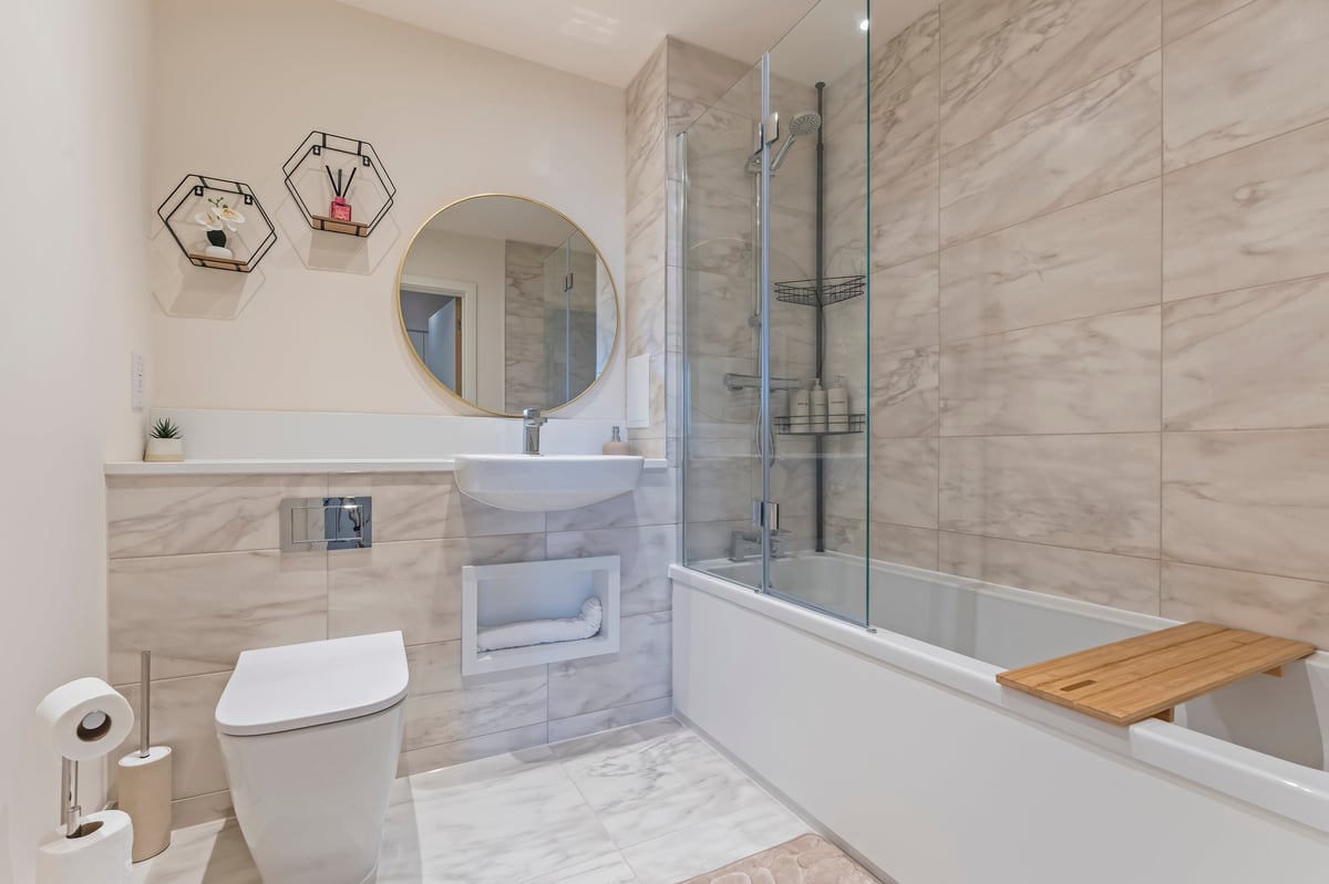 Bathroom at Wedgewood with marble tile walls, a round mirror above a floating vanity, a white toilet, a glass-enclosed shower over a full-length bath and a wooden bath mat
