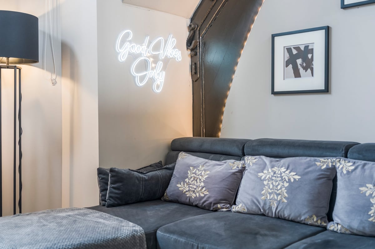 Bedroom at Athenaeum House with a dark blue L-shaped sofa, white neon Good Vibes Only sign, floor lamp and framed artwork on the wall