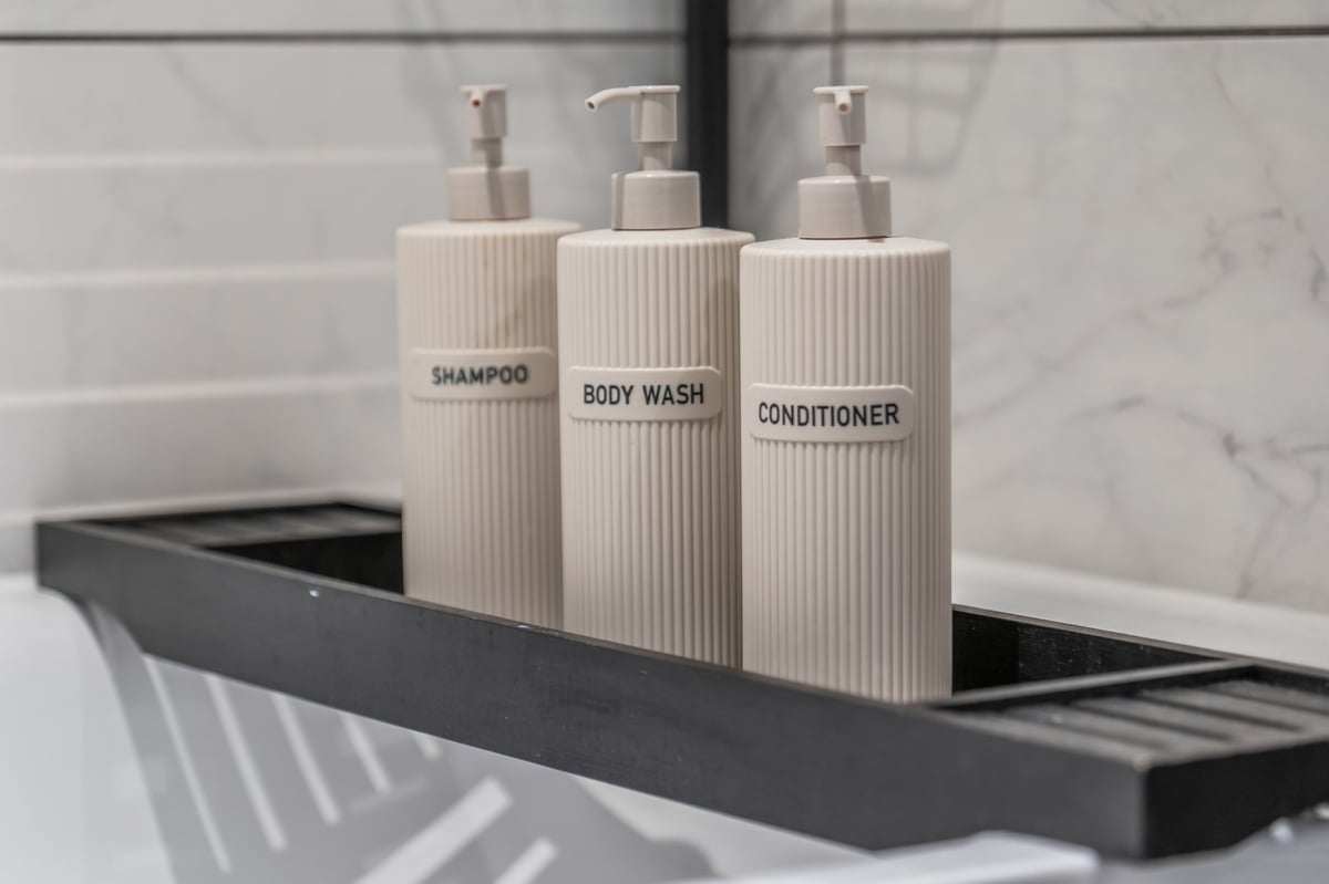 Three beige pump bottles labelled shampoo, body wash and conditioner arranged on a black shelf in the bathroom at the Aspire apartment