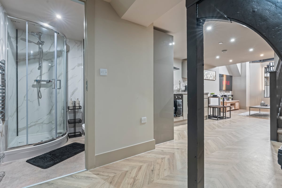 Modern interior at Athenaeum House showing the bathroom with a glass shower enclosure, cream walls, herringbone flooring and the open-concept living space beyond