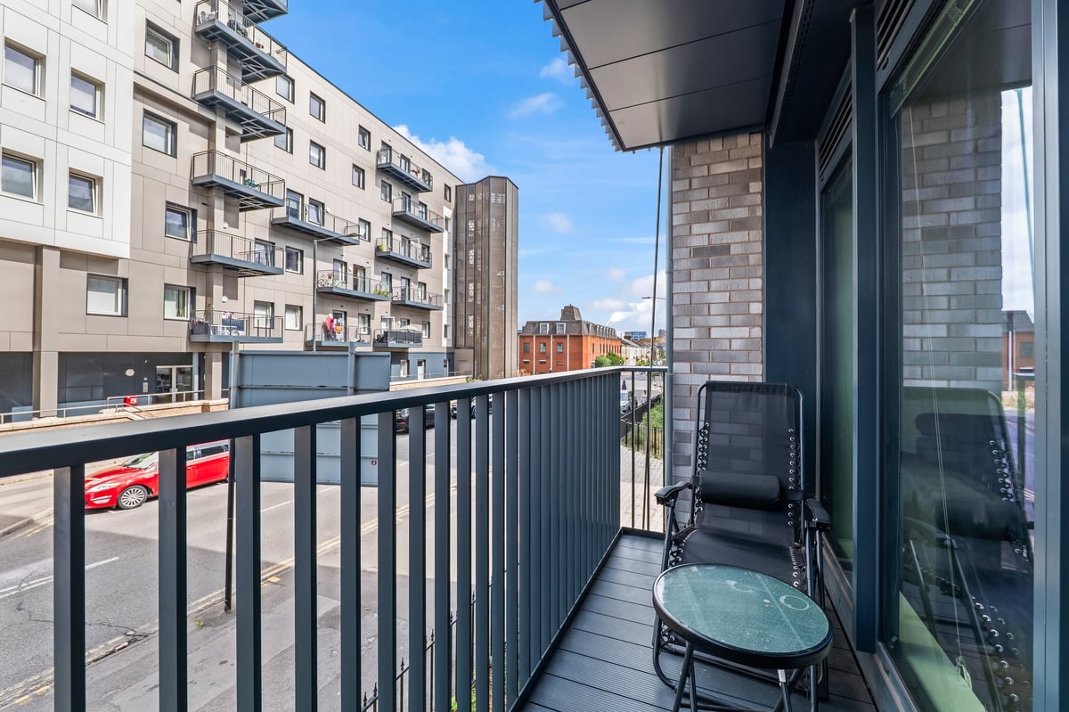 Balcony at the Aspire apartment with a black metal railing overlooking an urban street and apartment buildings on a sunny day, with a black chair and small round table