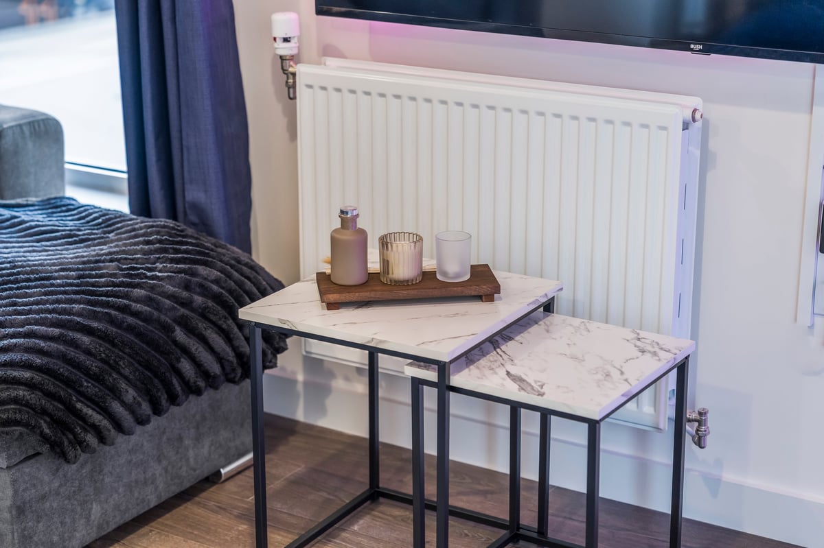 Modern bedroom at Wedgewood with a grey upholstered bed, nesting tables with decorative bottles, a white radiator and dark curtains