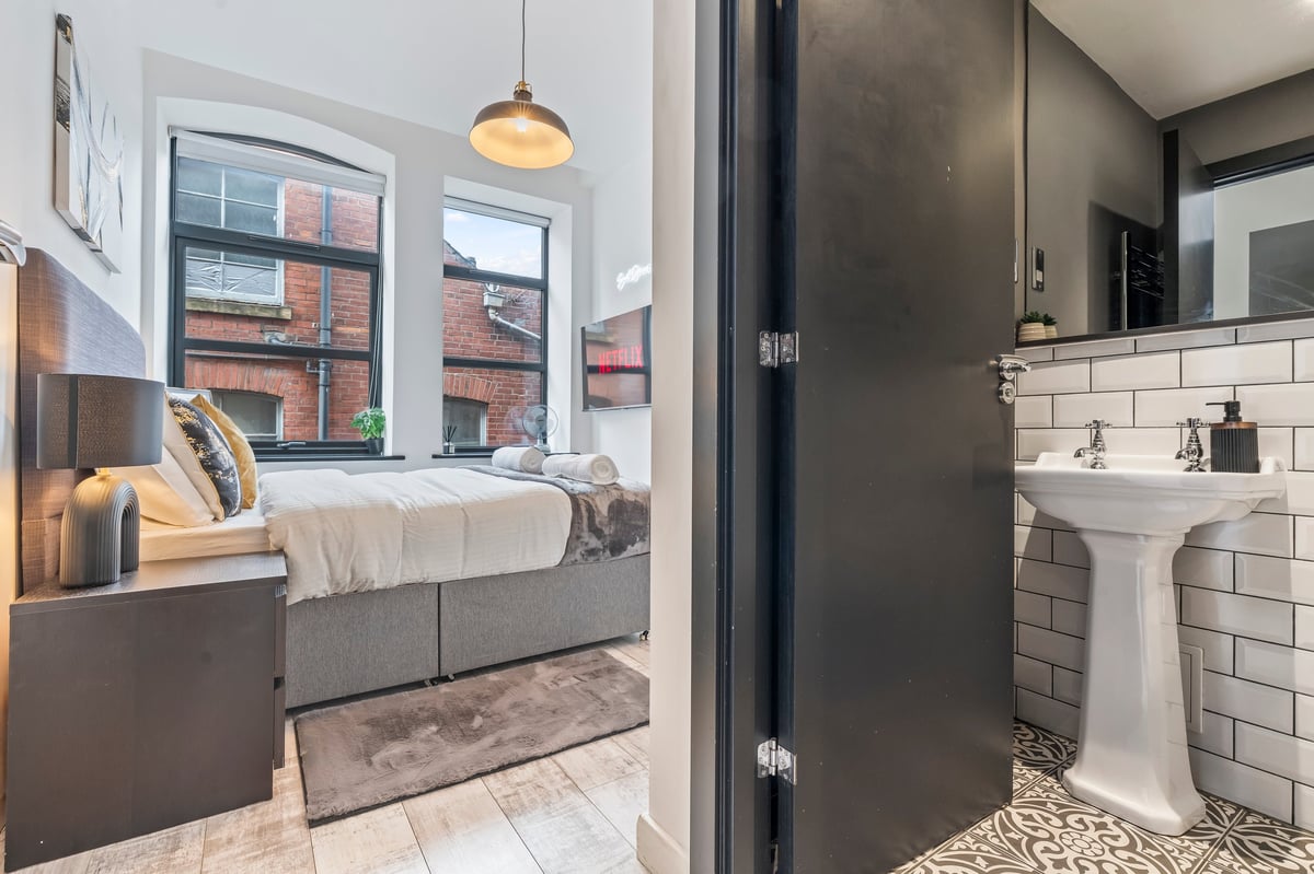 Split view of the bedroom and bathroom at The Calls showing the grey bed, pendant light and pedestal sink