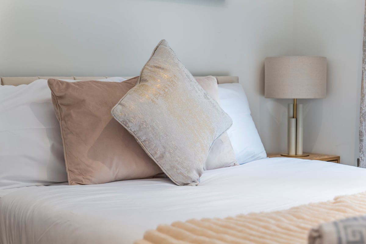 Bed at the Aspire apartment with decorative pillows in neutral tones, a beige bedside lamp and white linens in a minimalist bedroom