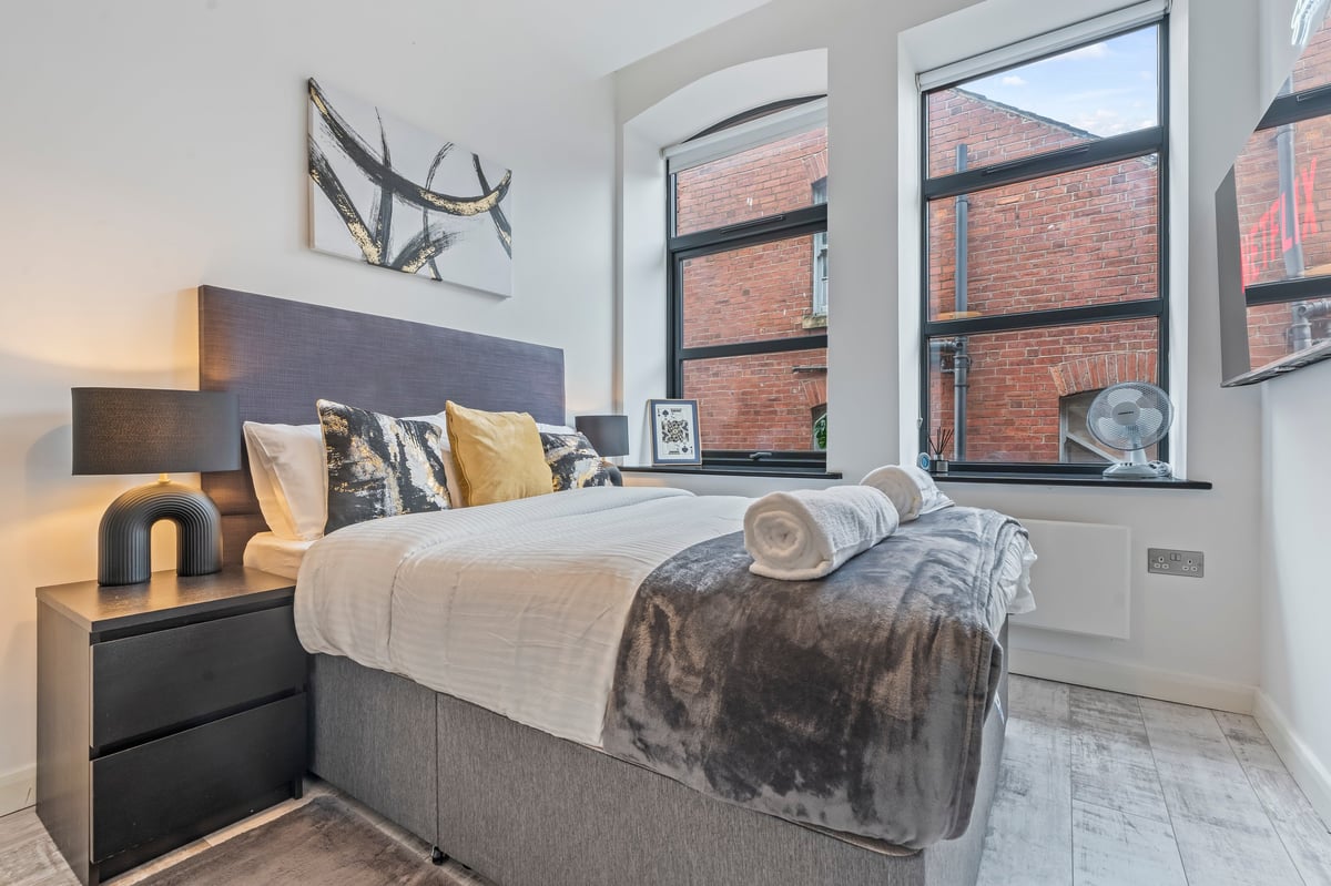 Bedroom at The Calls with a grey bed, yellow and white cushions and large windows looking out to brick buildings