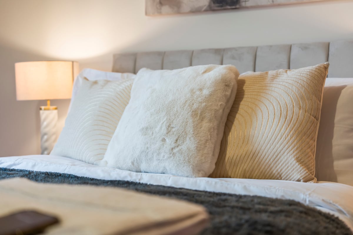 Bedroom at Wedgewood with layered decorative pillows in white and gold tones against a padded headboard with a bedside lamp