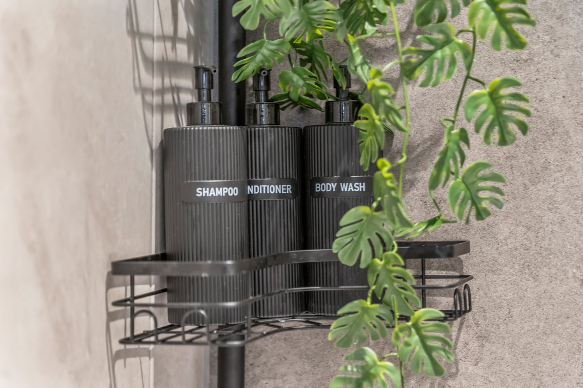 Black shower caddy holding three labelled bottles of shampoo, conditioner and body wash at the Aspire apartment, mounted on a grey wall with climbing green monstera vines