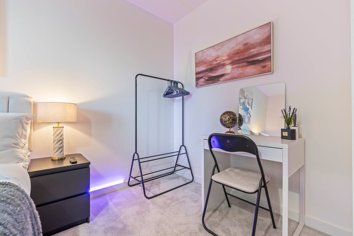 Corner of the bedroom at Wedgewood with a white desk, a black folding chair, a clothes rack and a nightstand with a lit lamp and purple accent lighting