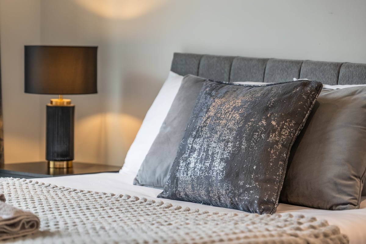 Bedroom at the Aspire apartment with a grey upholstered headboard, decorative pillows, white bedding and a bedside lamp with a dark shade on the nightstand