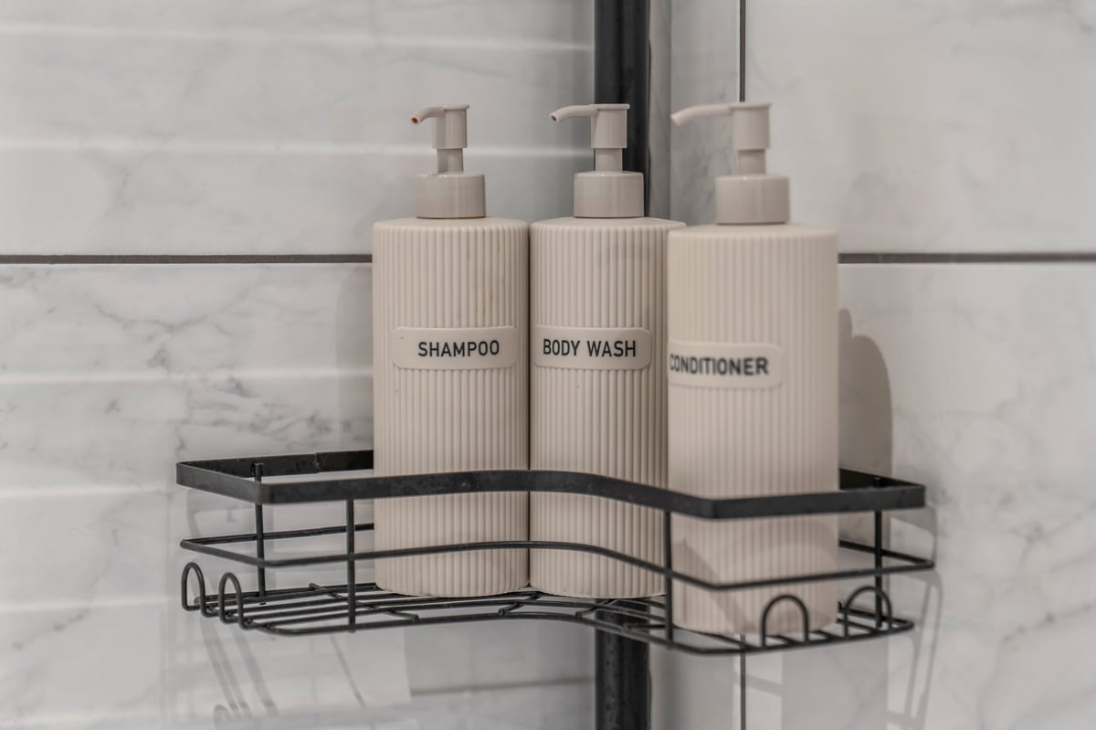 Three cream-coloured pump bottles labelled shampoo, body wash and conditioner at the Aspire apartment, arranged in a black metal shower caddy mounted on white tile