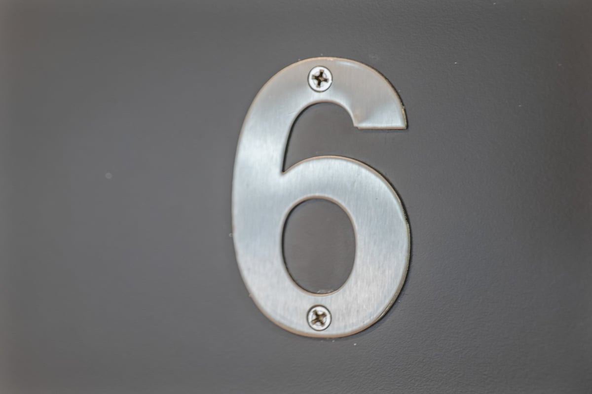 Metal number 6 with a brushed silver finish and grey cutouts at the Aspire apartment, mounted with screws on a dark background