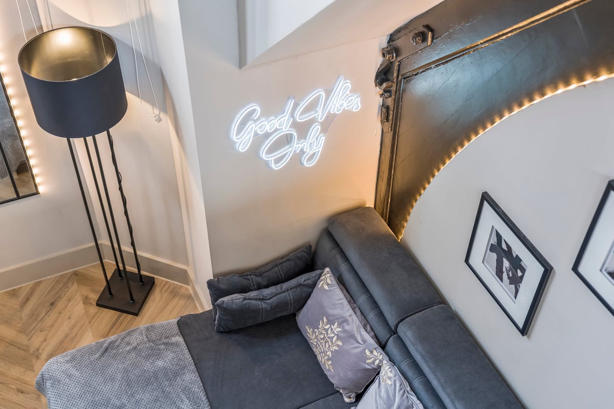 Cozy bedroom corner at Athenaeum House with a neon Good Vibes Only sign, a black floor lamp, the bed with pillows and framed wall art