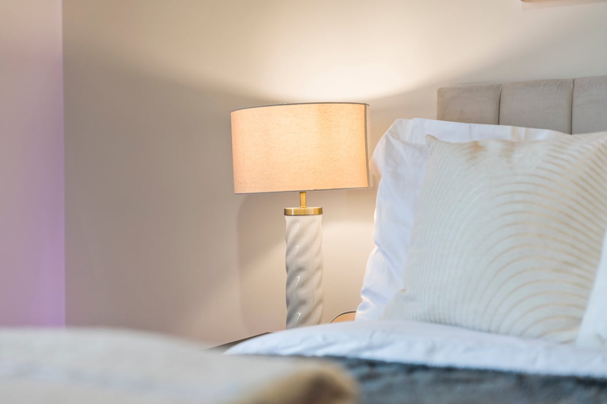 Bedroom nightstand at Wedgewood with an illuminated lamp and white pillows on the bed against a neutral wall