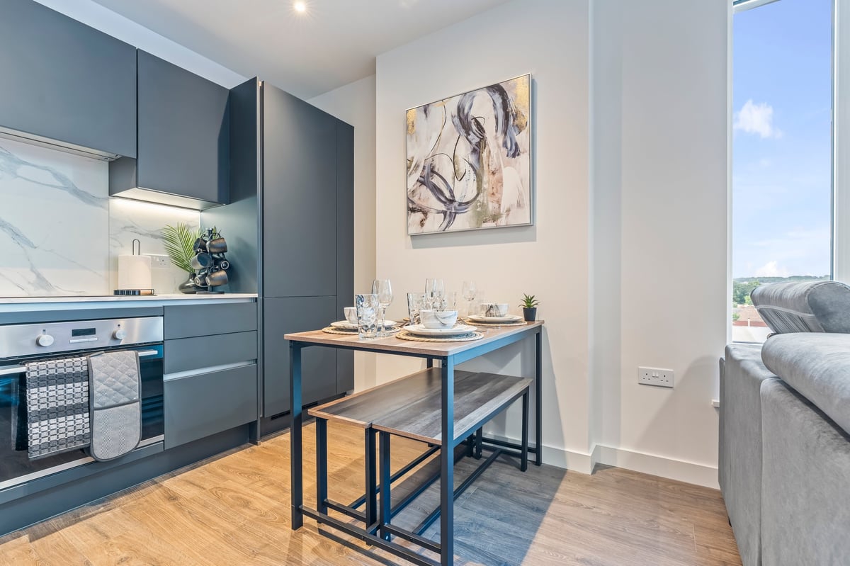 Compact kitchen at Wedgewood with dark blue cabinetry, a small dining table and the adjoining living area with a grey sofa