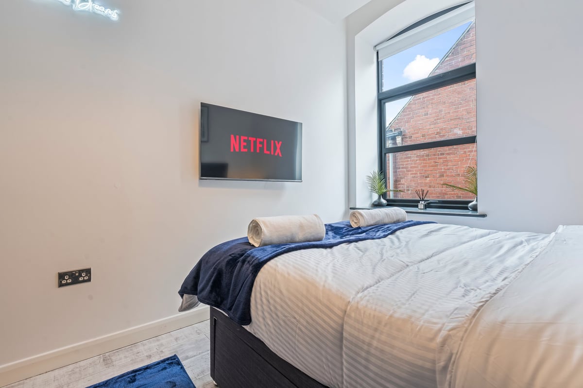 Bedroom at The Calls with a wall-mounted television above the bed, white and blue bedding and a brick exterior view