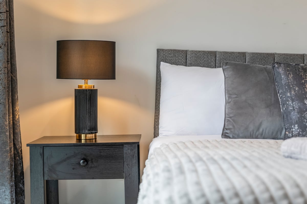 Bedroom nightstand at the Aspire apartment with a dark table lamp and dark headboard behind white and grey pillows on the bed