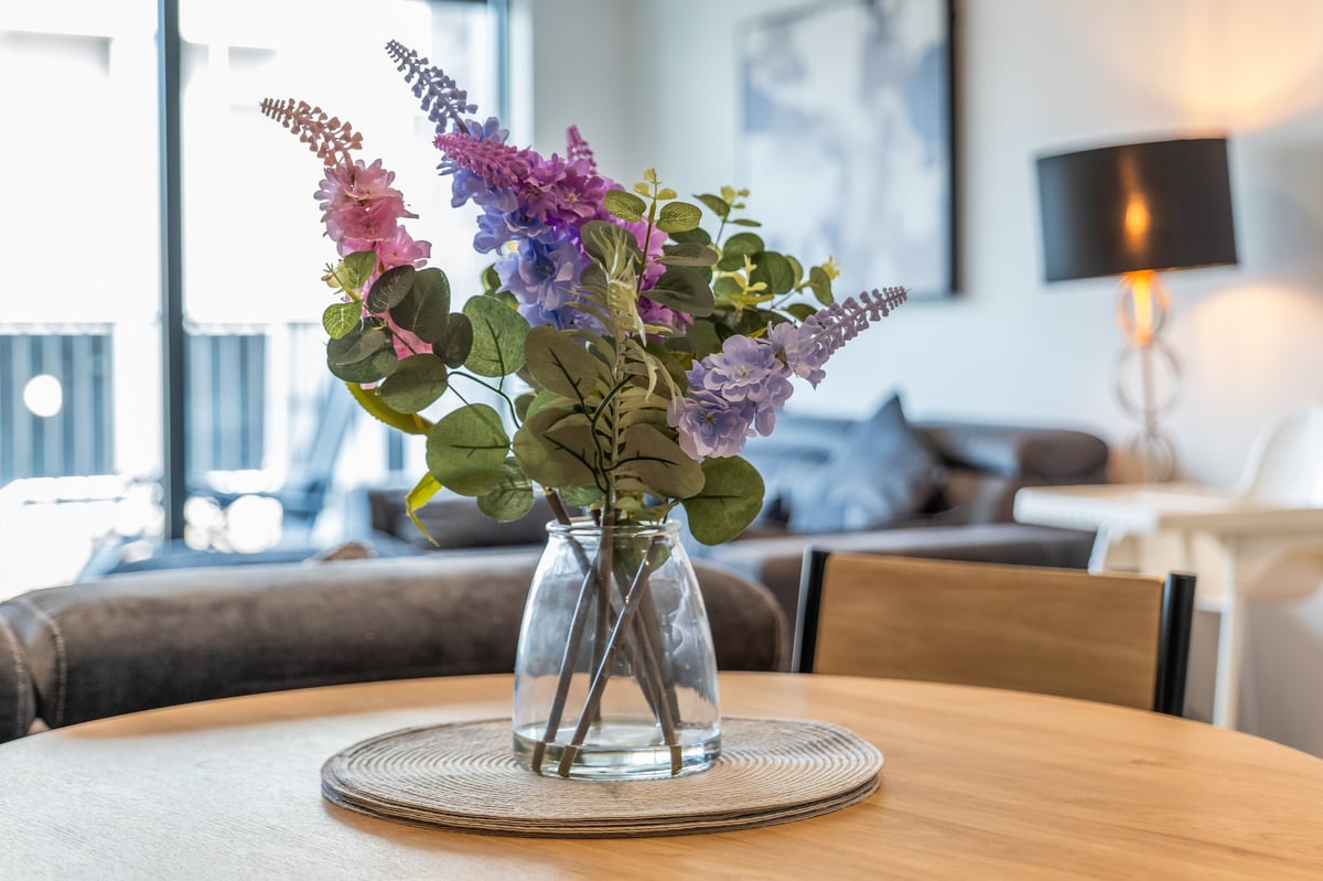 Glass vase of purple and lavender flowers on a wooden table at the Aspire apartment, with the modern living room beyond