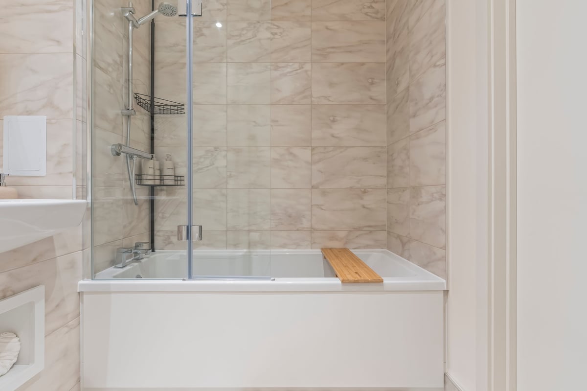 Bathroom at Wedgewood with a glass shower enclosure over a white bathtub, marble tile walls and a wooden bath caddy