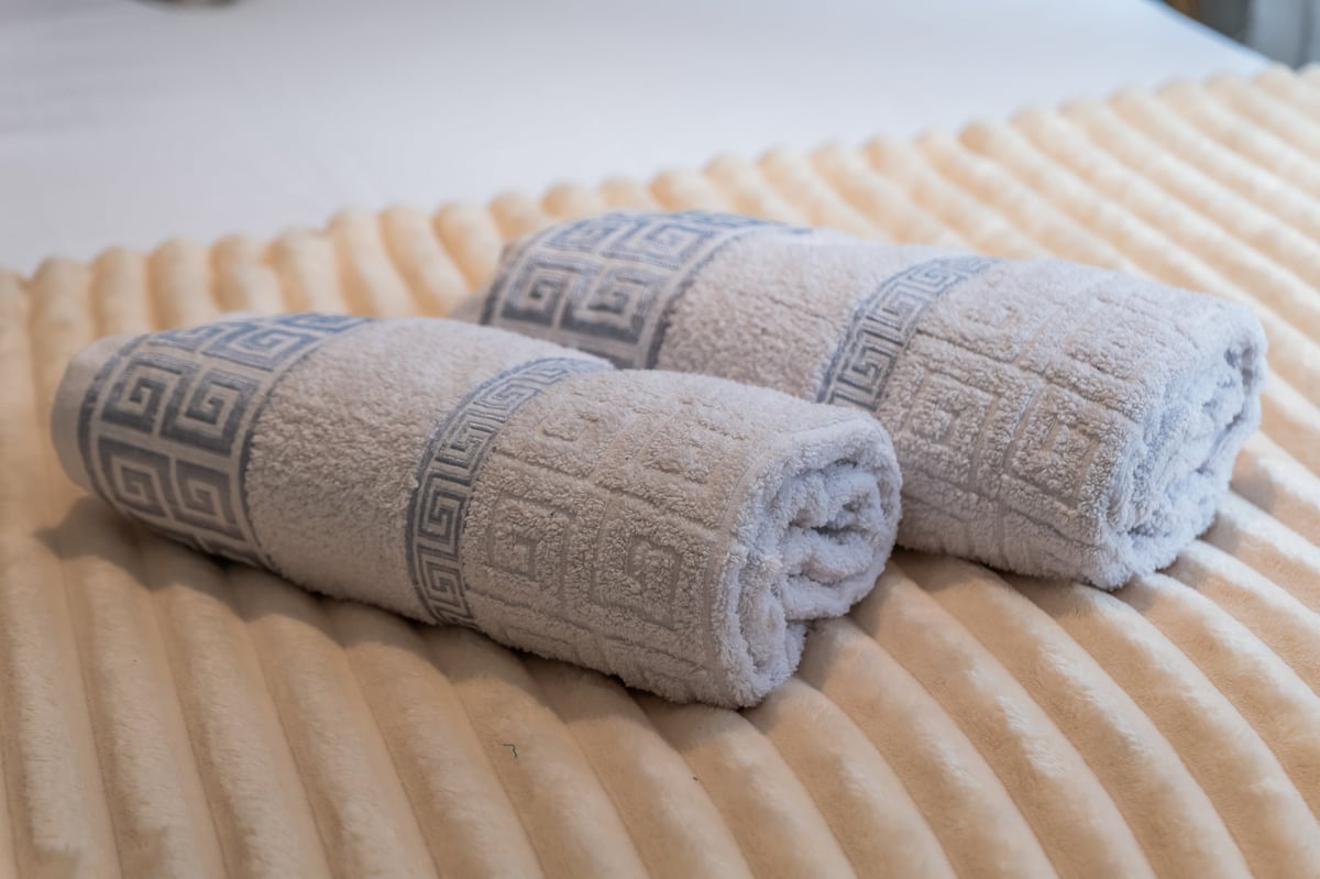 Two rolled beige towels with Greek-key pattern bands at the Aspire apartment, displayed on a textured cream bedspread