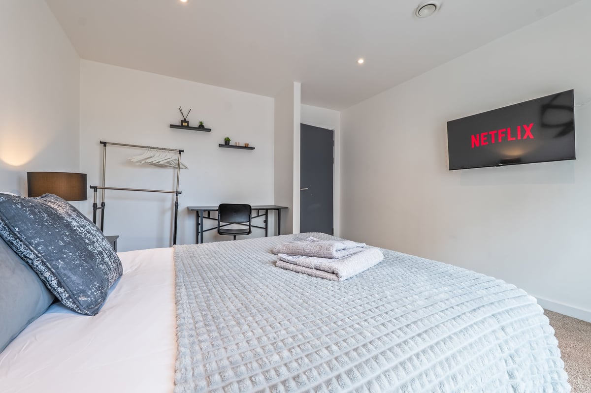 Bedroom at the Aspire apartment with white bedding, Netflix displayed on a wall-mounted television, shelving and minimalist decor