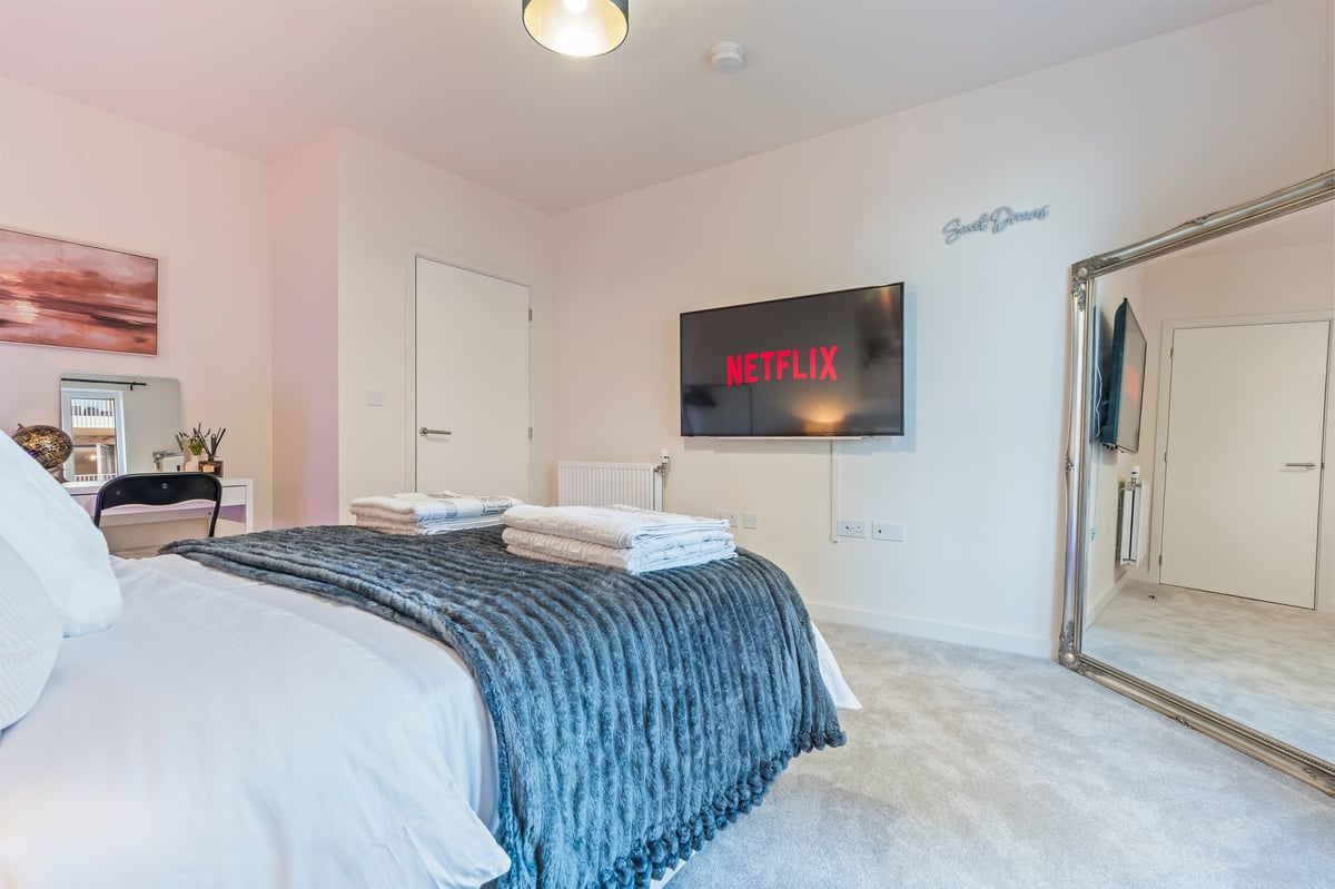 Bedroom at Wedgewood with a white bed, blue blanket, stacked folded towels, a wall-mounted television displaying the Netflix home screen, a full-length mirrored wardrobe and a reading nook with a chair and side table