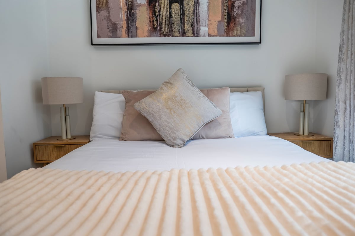 Neatly made bed at the Aspire apartment with striped cream bedding, decorative pillows in neutral tones, wooden nightstands with table lamps, and a framed abstract artwork above the headboard