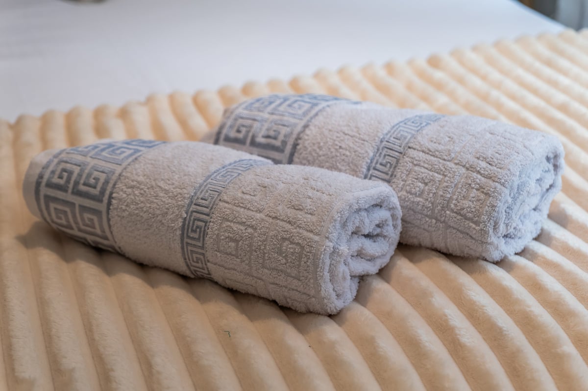 Three rolled beige towels with geometric Greek-key pattern bands laid on a cream-coloured quilted bedspread at the Aspire apartment