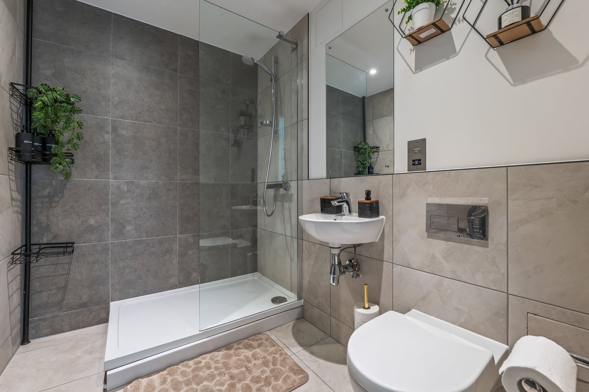Bathroom at the Aspire apartment with a walk-in shower, white toilet, beige tiles, glass enclosure and hanging plants above the sink