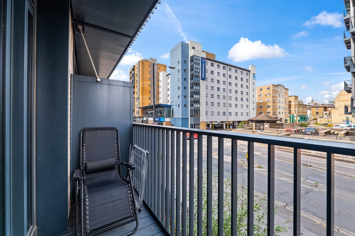 Narrow balcony at the Aspire apartment with a black reclining chair overlooking the urban cityscape of residential and office buildings under a blue sky