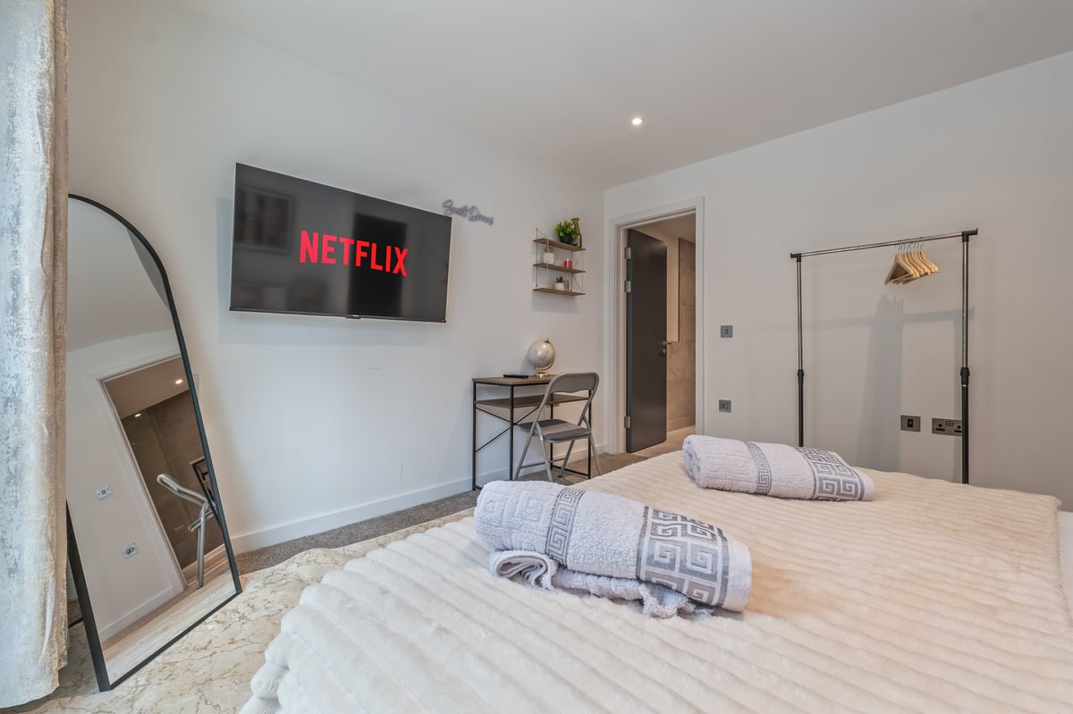 Bedroom at the Aspire apartment with a wall-mounted Netflix television, a queen bed with white bedding, a round mirror and a glass shower enclosure in a minimalist layout