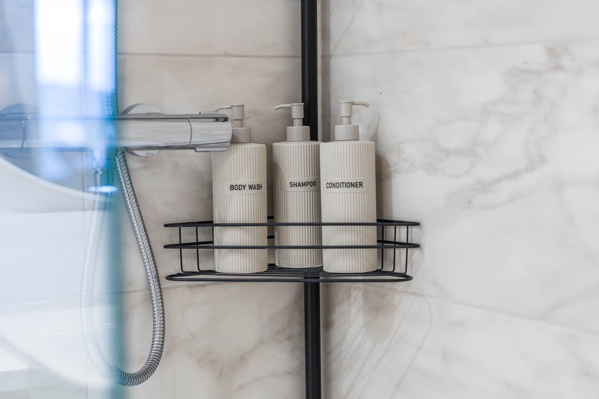Black wire shower caddy at Wedgewood with three labelled bottles of body wash, shampoo and conditioner on a marble tile wall
